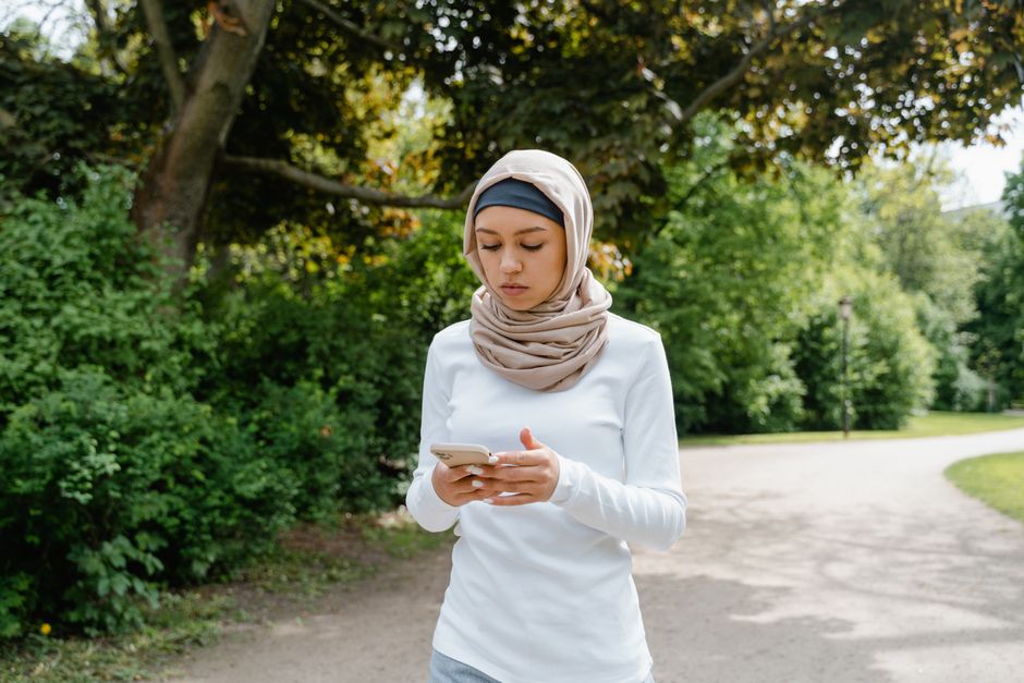 Muslim woman wearing hijab walking in a park, using cellphone, surrounded by green trees.